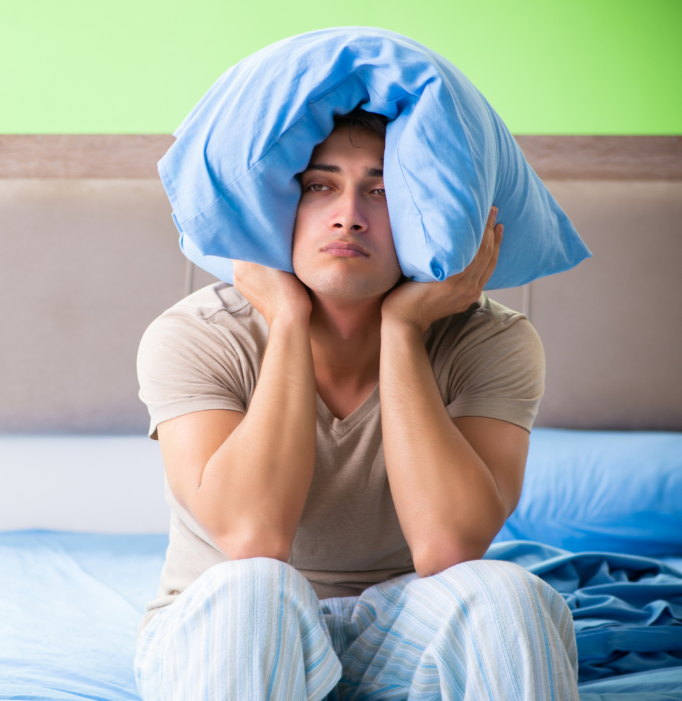 Struggling with sleep disorders, shown by his action of putting a pillow over his head, highlighting the challenges and frustrations associated with disrupted sleep patterns.